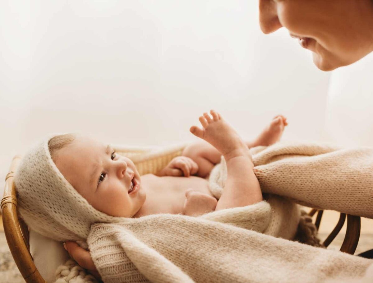 Ruua Hamdan Photography (UK): A baby wrapped in a cream-colored blanket lies in a basket, looking up at an adult's smiling face.