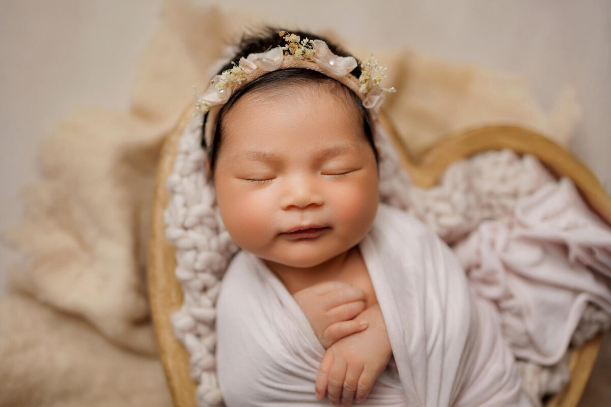 A peaceful newborn baby girl sleeps swaddled in a white blanket, adorned with a delicate floral headband. She rests in a heart-shaped basket lined with soft, creamy fabric.