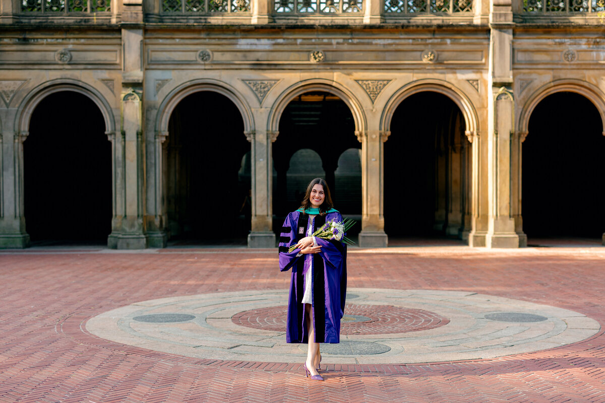 NYU graduate smiling while holding purple flowers and wearing purple gown at Central Park.