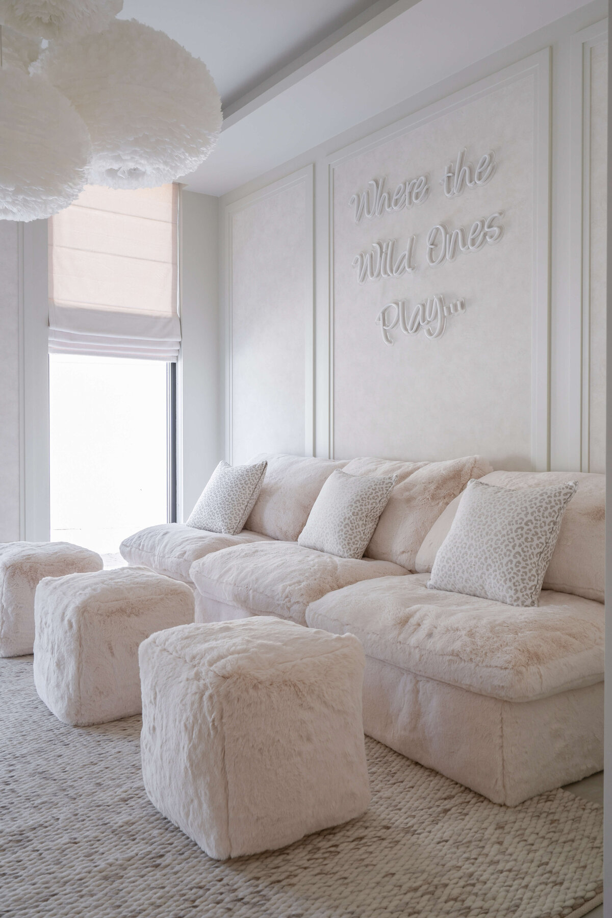 Spacious playroom with a light, airy aesthetic. There is a large, plush tan couch with three fuzzy pouf ottomans. Behind the couch is a neon  sign reading 'Where the Wild Ones Play...'. Above are four white, feathery spherical pendant chandeliers.