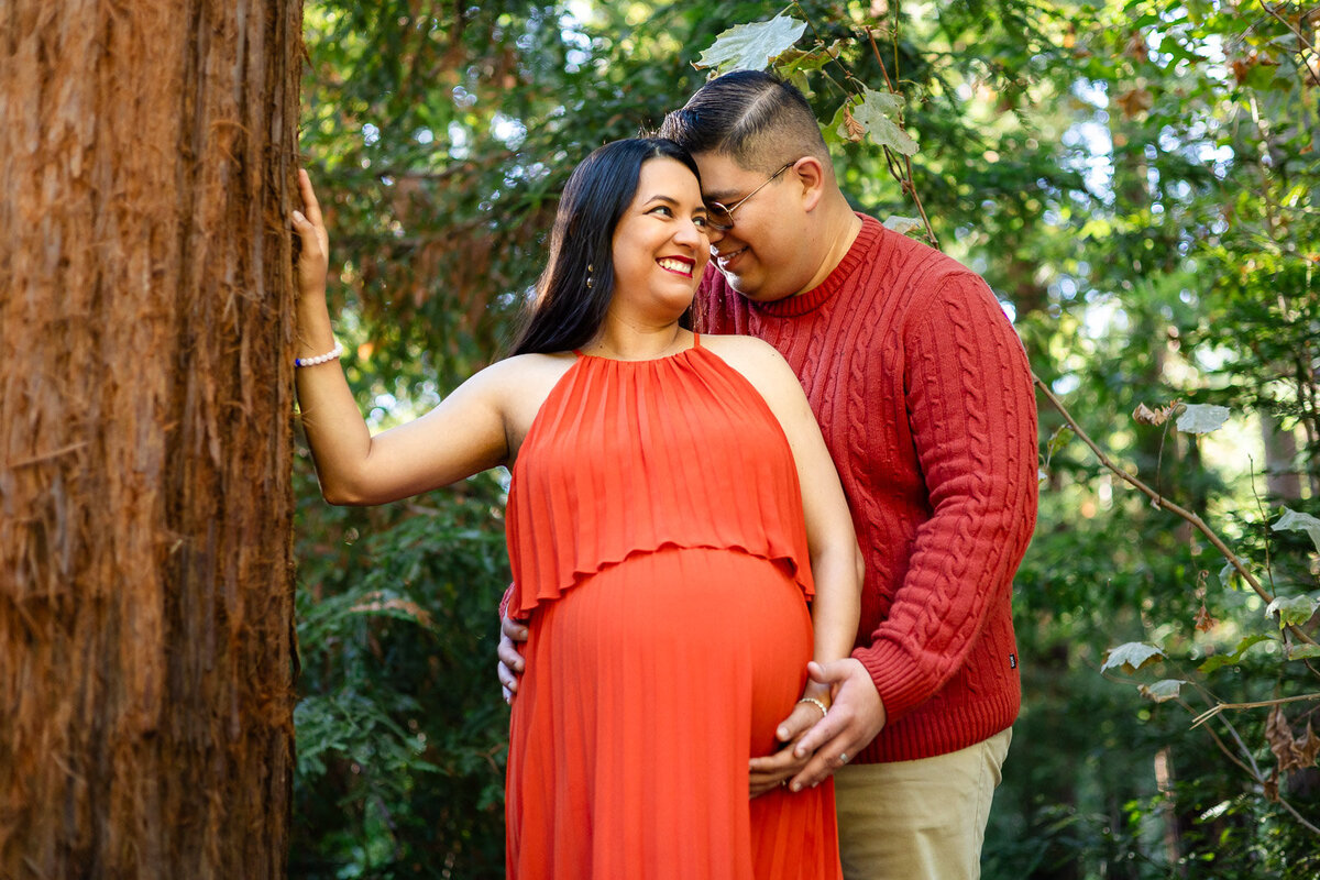 Pregnant couple embracing and smiling in dappled light by a large redwood tree – Ellobelle Photography