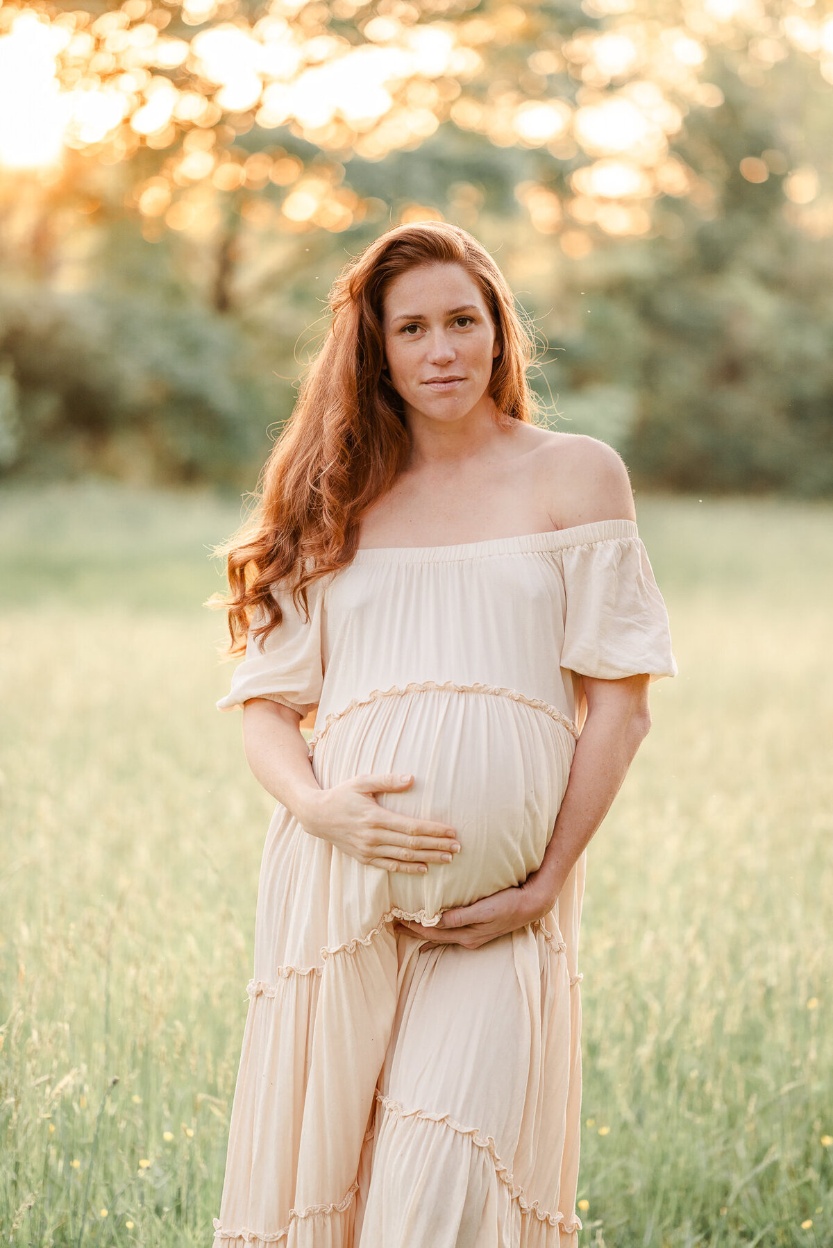 An expectant mother holds onto her belly while looking at the camera during her Chesapeake maternity session. She is wearing a long off-white dress and standing in a grassy field.