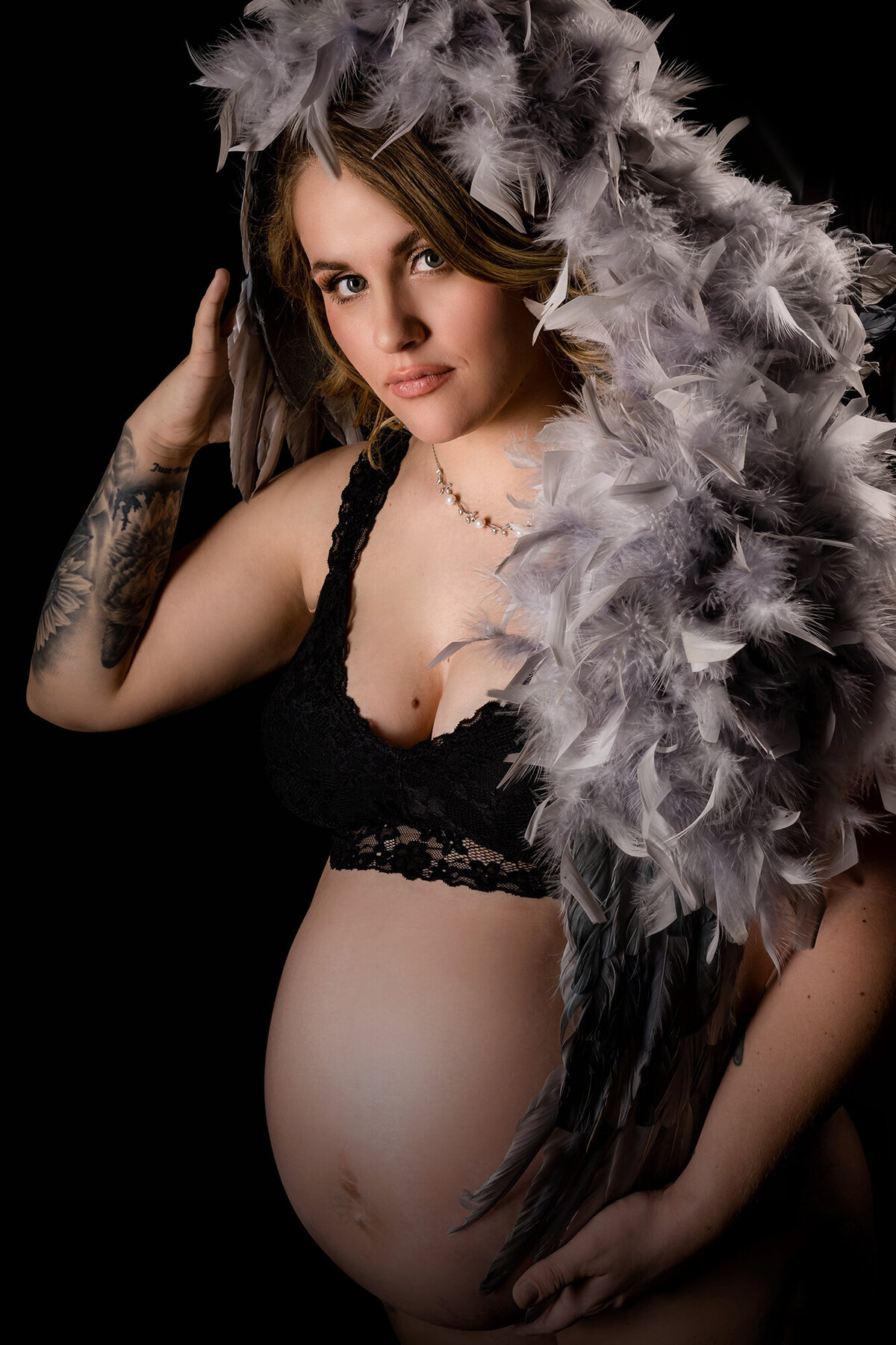 A pregnant woman stands in a black lace bra and grey feather headdress