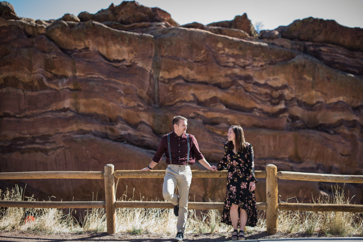 A wide angle shot of an engaged couple standing in front of a fence at Red Rocks in Colorado.