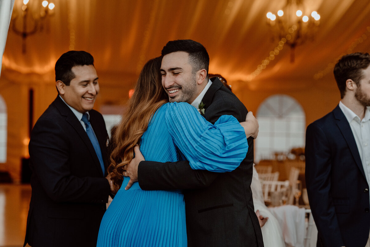 Groom warmly embraces a guest in a bright blue dress during the wedding reception, capturing a candid moment of joy and celebration. The elegant indoor setting, with warm lighting and chandeliers, enhances the festive atmosphere as guests interact and share smiles.