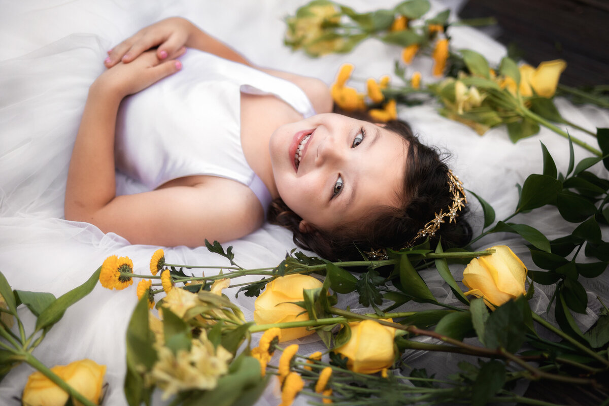 Young girl lying on the ground in a fancy white dress.  She has very dark hair.  She is looking up towards the camera and her hands are crossed at her waist.  She is surrounded by yellow roses.