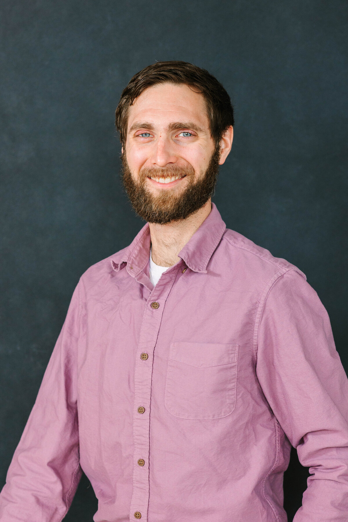 A bearded man with short brown hair smiles at the camera, wearing a mauve button-up shirt. He stands against a dark, textured backdrop.