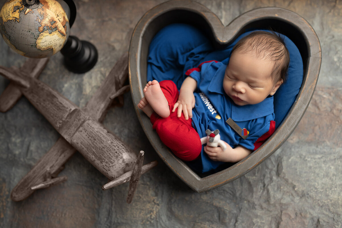 Close-up photo of a newborn in a Southwest Airlines uniform with a name tag that reads “Koa,” resting in a heart-shaped bowl – baby photographer in Fort Worth