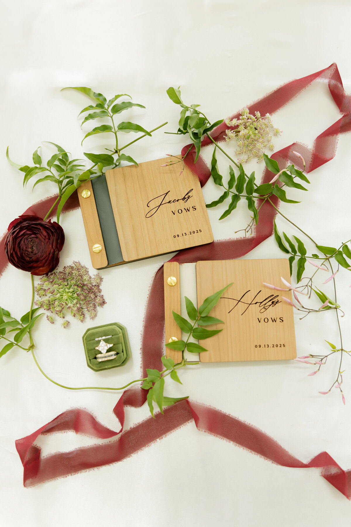 A styled flatlay of Jacob and Holly’s engraved wooden wedding vow books surrounded by greenery, mauve ribbon, wildflowers, and a green velvet ring box featuring a diamond engagement ring. The natural, organic styling evokes a romantic garden wedding theme.
