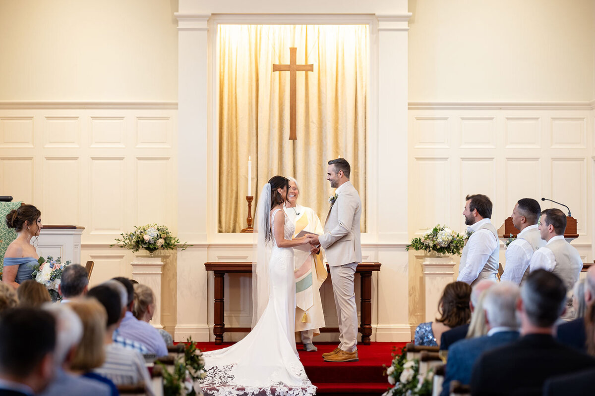A bride and groom smiling at each other inside a church, with a large cross in the background and guests seated on either side.
