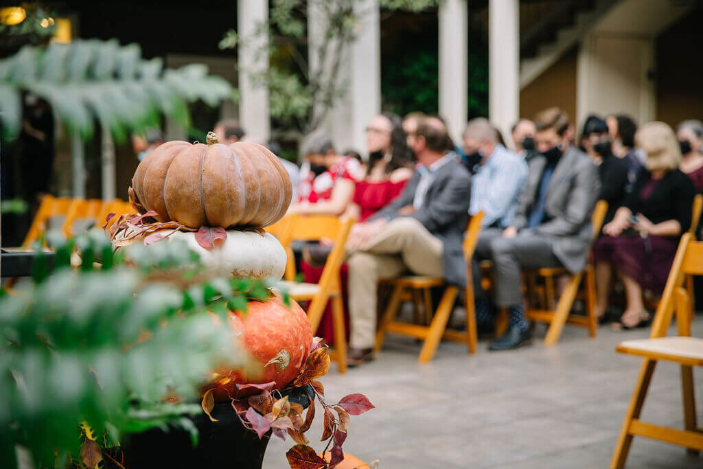 A stack of orange, white, and brown pumpkins with autumn leaves in the foreground; people in formal attire are seated on wooden chairs in the blurred background, suggesting a fall-themed event or ceremony.