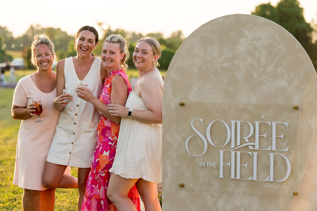 the four female organizers of Soiree in the Field holding drinks and smiling at the camera.  Captured by Ottawa Event Photographer JEMMAN Photography COMMERCIAL