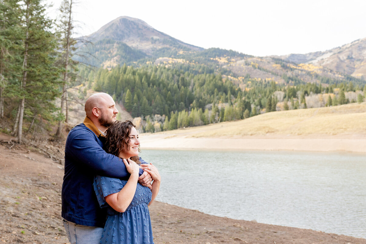 A couple embraces lovingly by the serene waters of Tibble Fork Reservoir in Utah, with a stunning mountain backdrop showcasing autumn foliage. The man in a navy blue jacket holds the woman in a blue dress as they share a quiet moment together.
