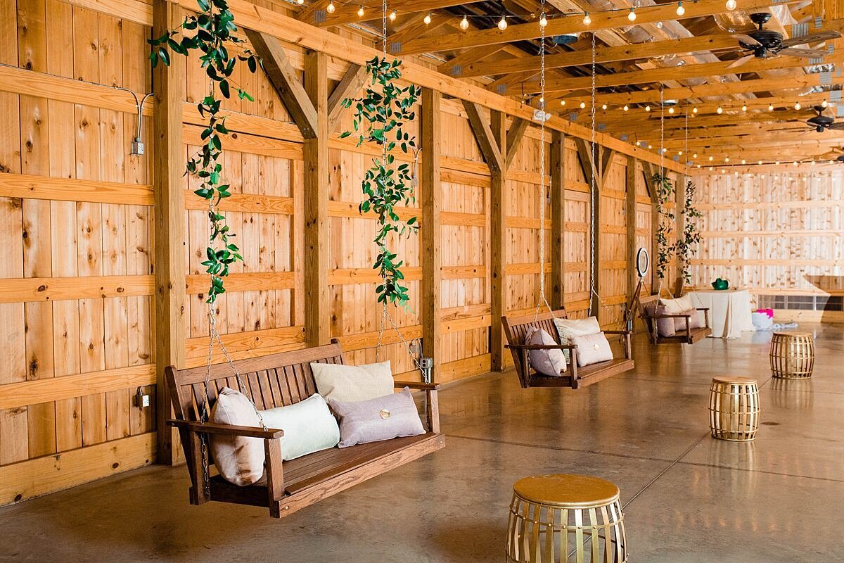 Three wood porch swings hang from the ceiling of the cedar barn at Saddle Woods Farm. The swings are decorated with Italian ruscus vine, ivory and tan throw pillows with gold moroccan drum tables.