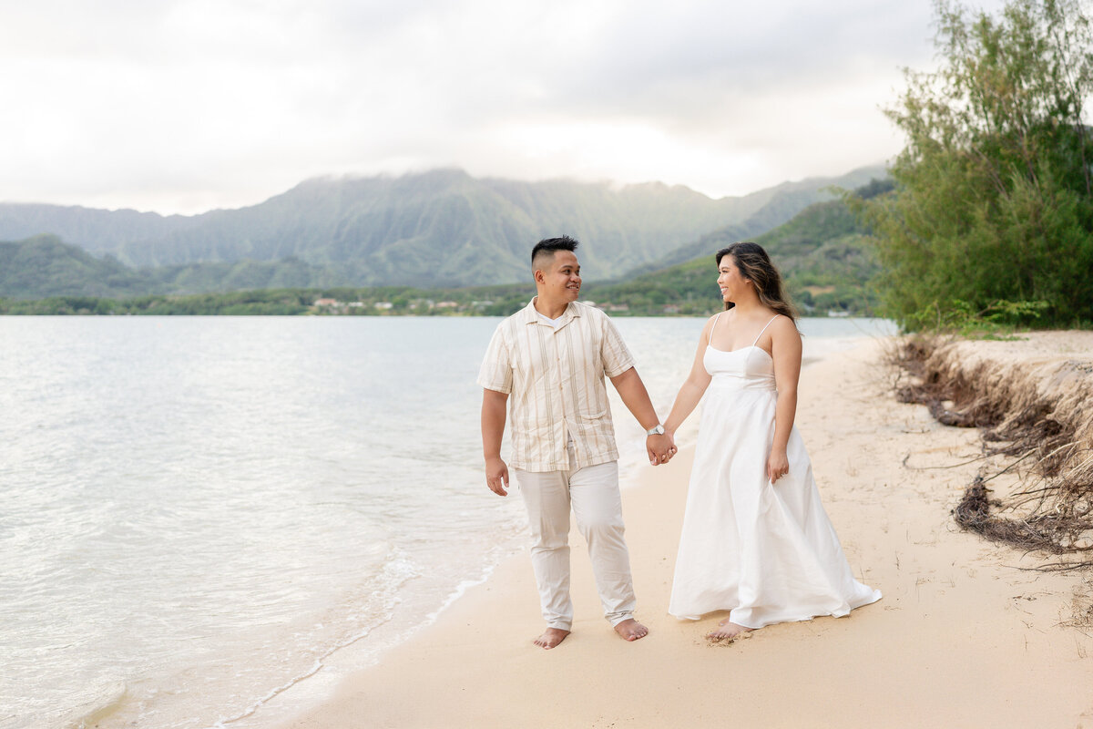 Oahu Hawaii Elopement Adventurous Asian couple at Secret Island Beach during golden sunset with mountains and ocean waves holding hands walking.