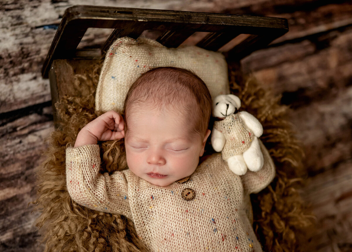A newborn baby sleeps in a beige knit onesie with a bear in a matching onesie on a tiny wooden bed in a Des Moines Newborn Photography studio