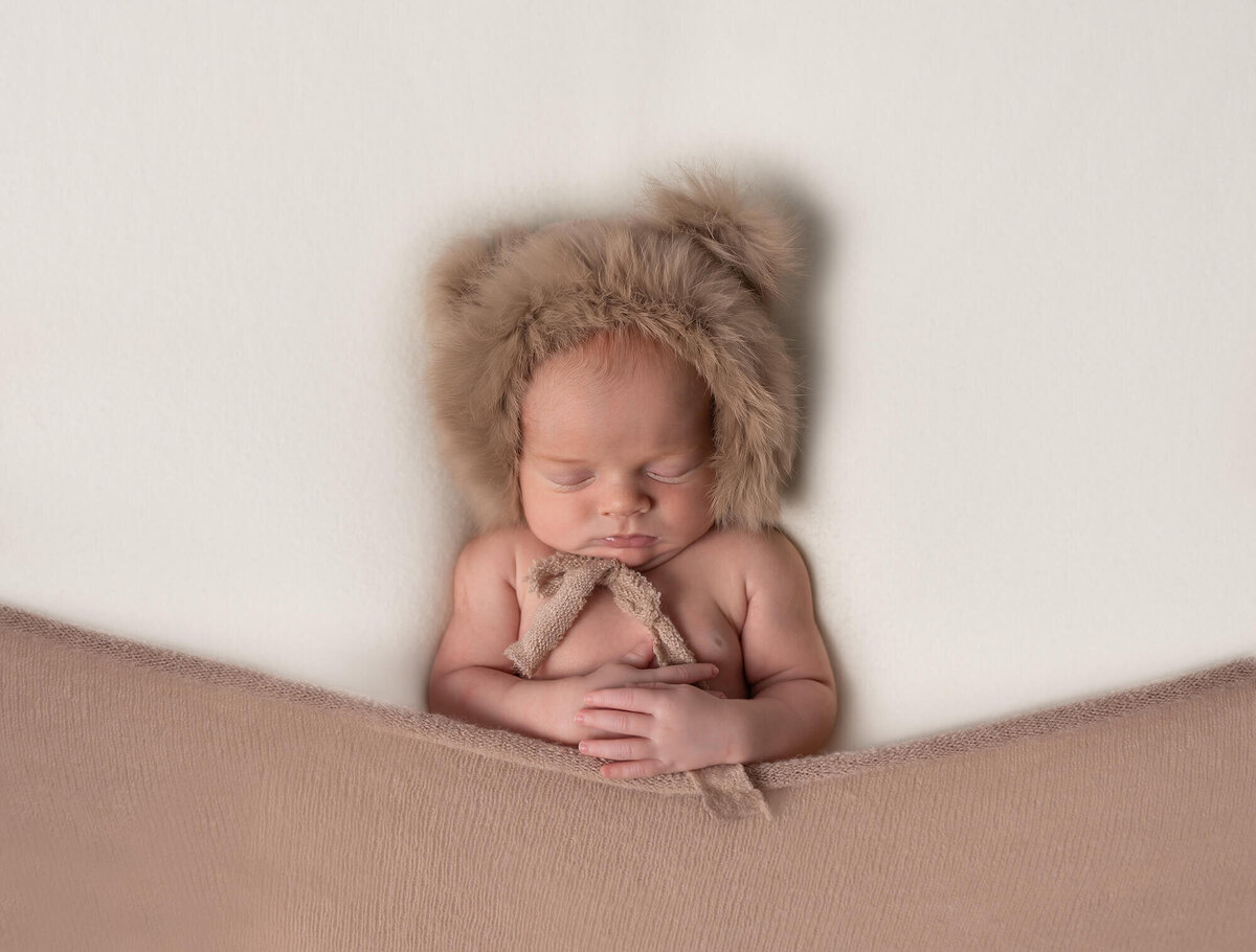 A newborn baby boy sleeps under a brown blanket with a fuzzy teddy bear hat on