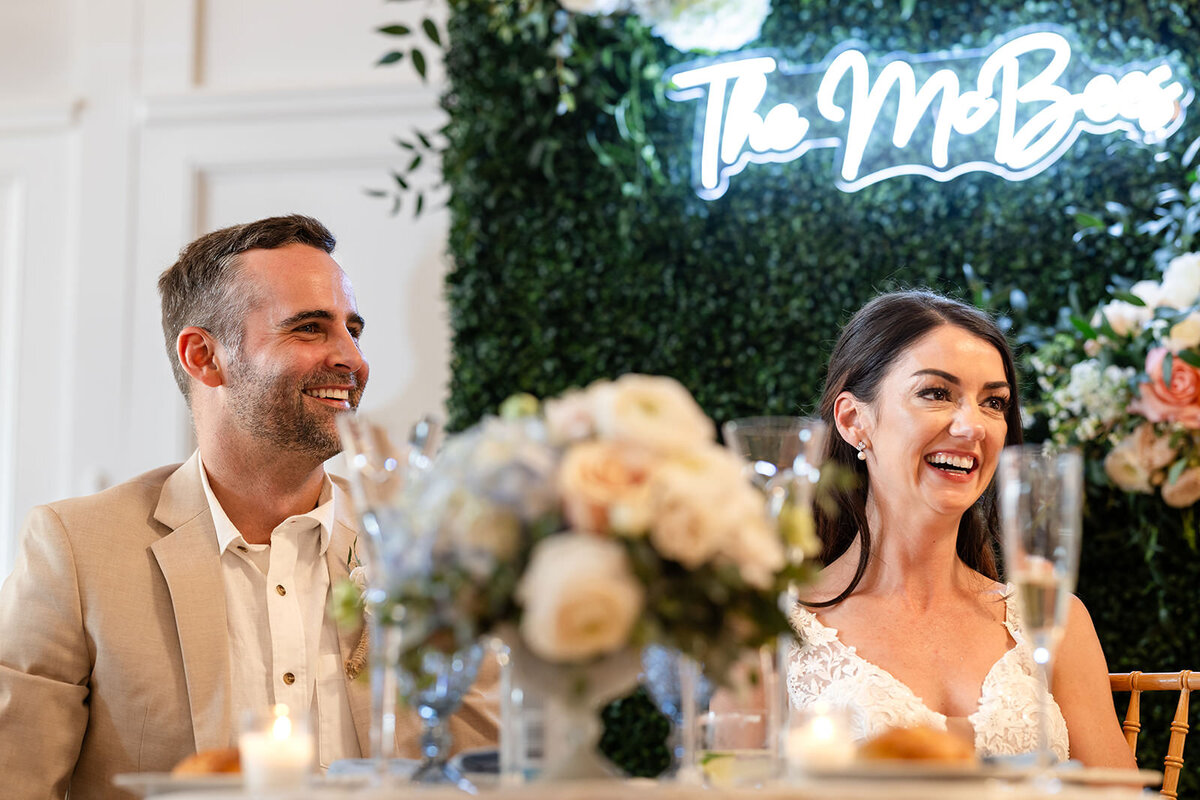 The bride and groom at their wedding table, smiling joyfully, with a neon sign "The McBee's" and floral arrangements in the background.