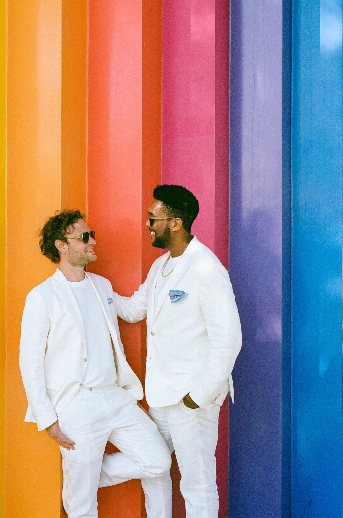 A couple smiling and leaning up against a rainbow wall.