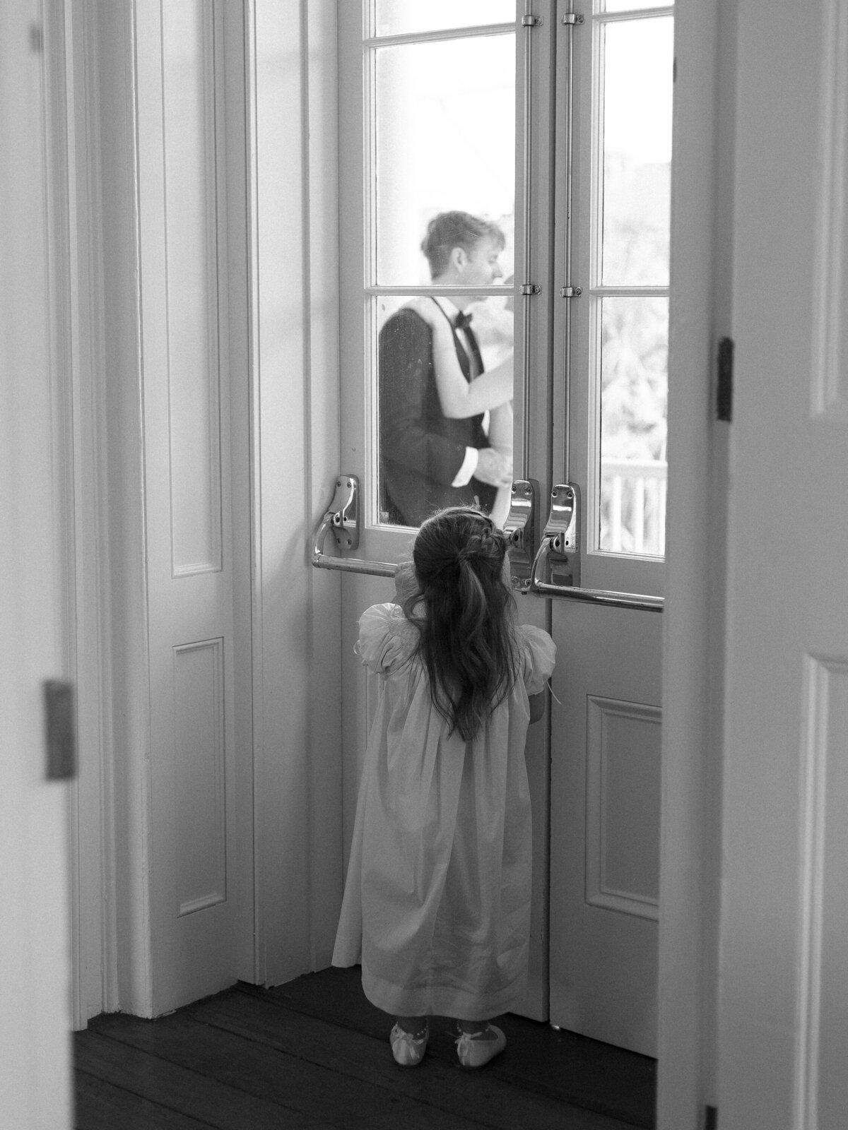 Flower girl looks through a window in a door at the bride and groom embracing