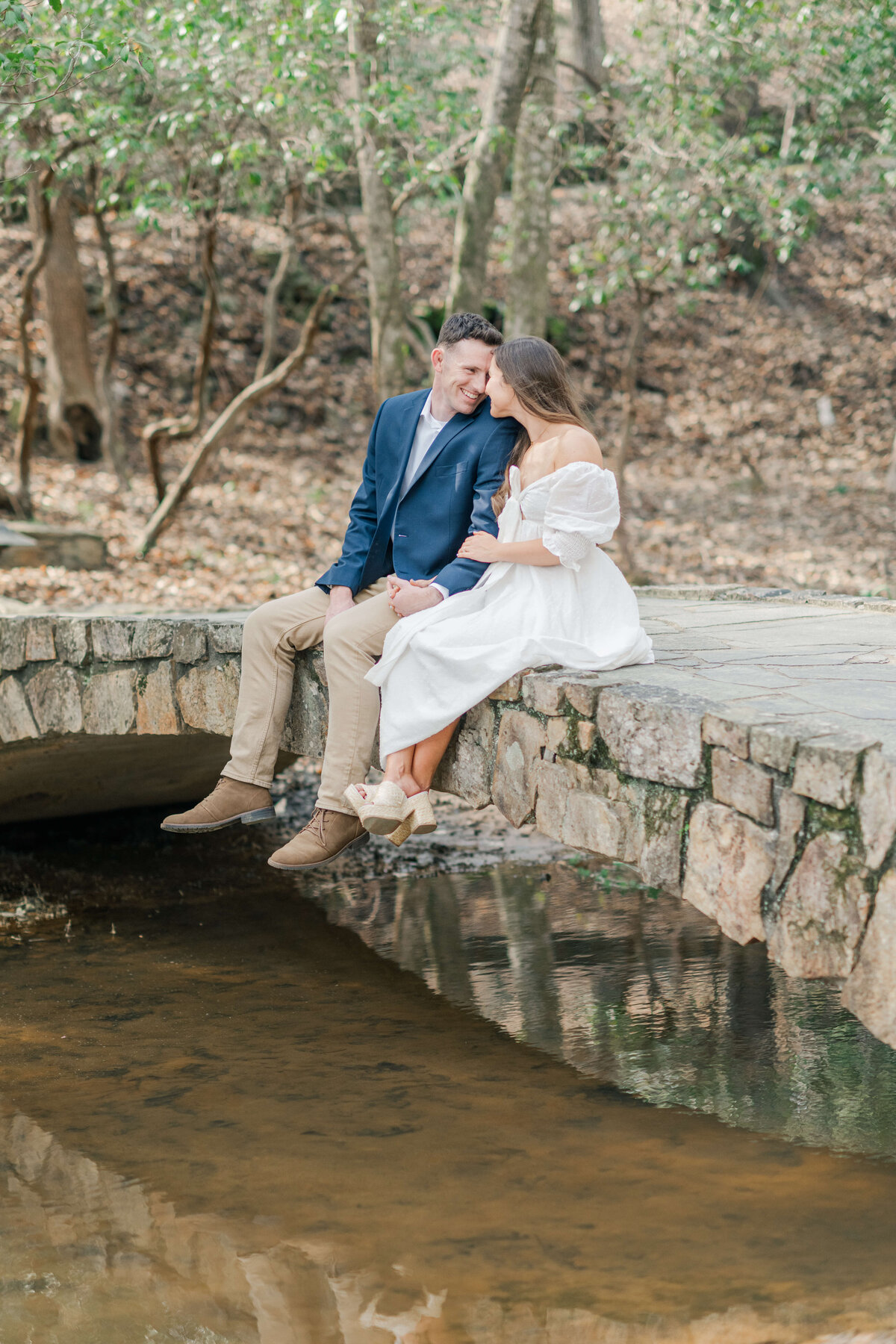 Fiances sit on a bridge above a stream looking at eachother smiling for enggaement photos