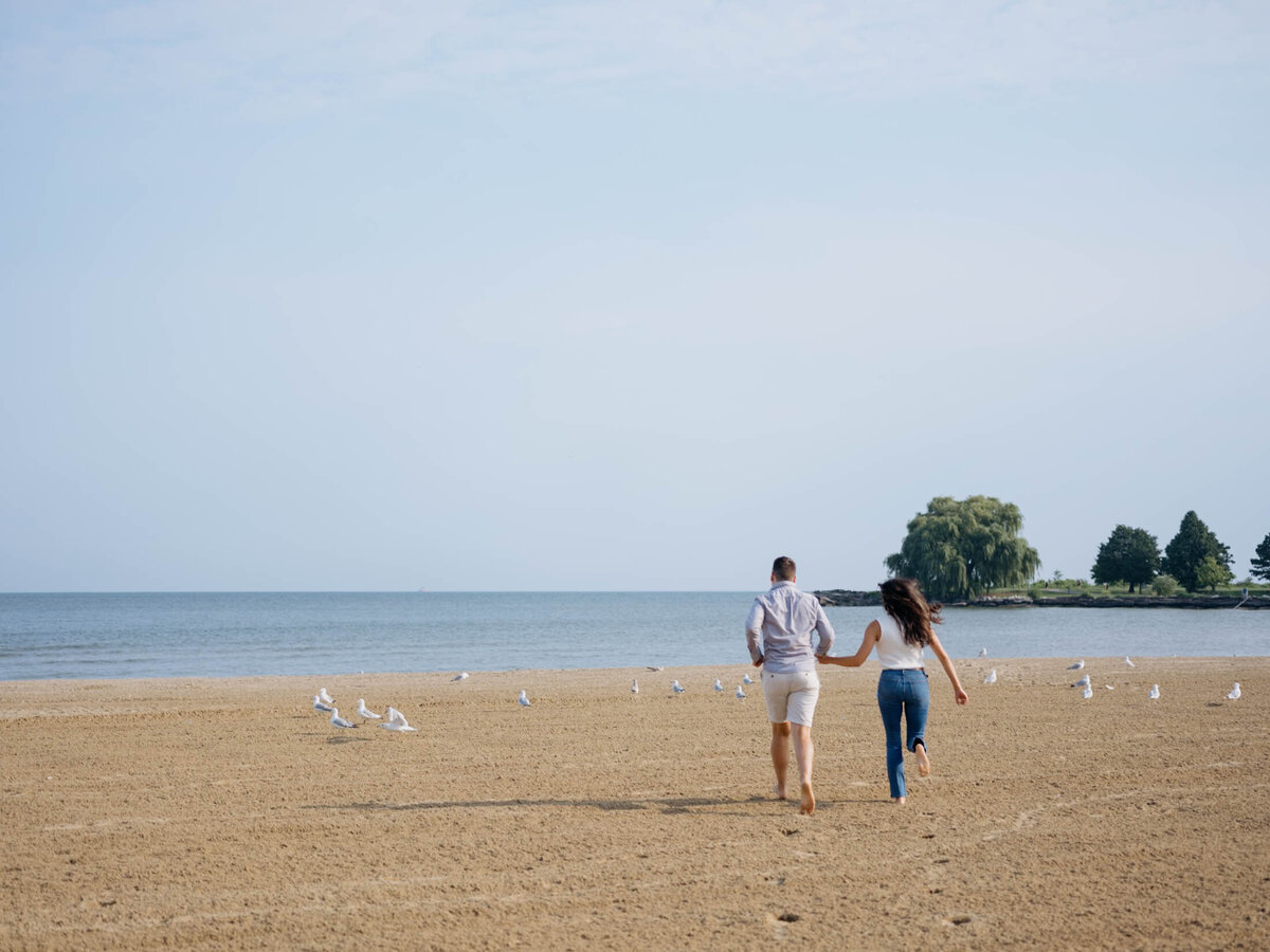 Engagement photo of a couple running hand in hand along a sandy beach in Cleveland, Ohio.