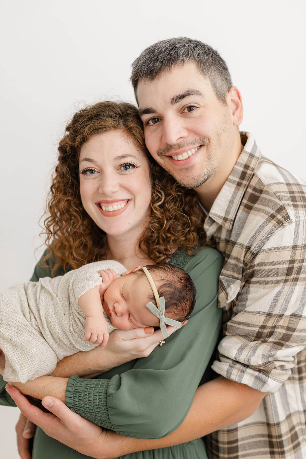 Mom in green dress holding sleeping newborn girl in her arms, dad is hugging her from behind. Dad is wearing a neutral plaid shirt. Mom and Dad are both smiling at the camera.