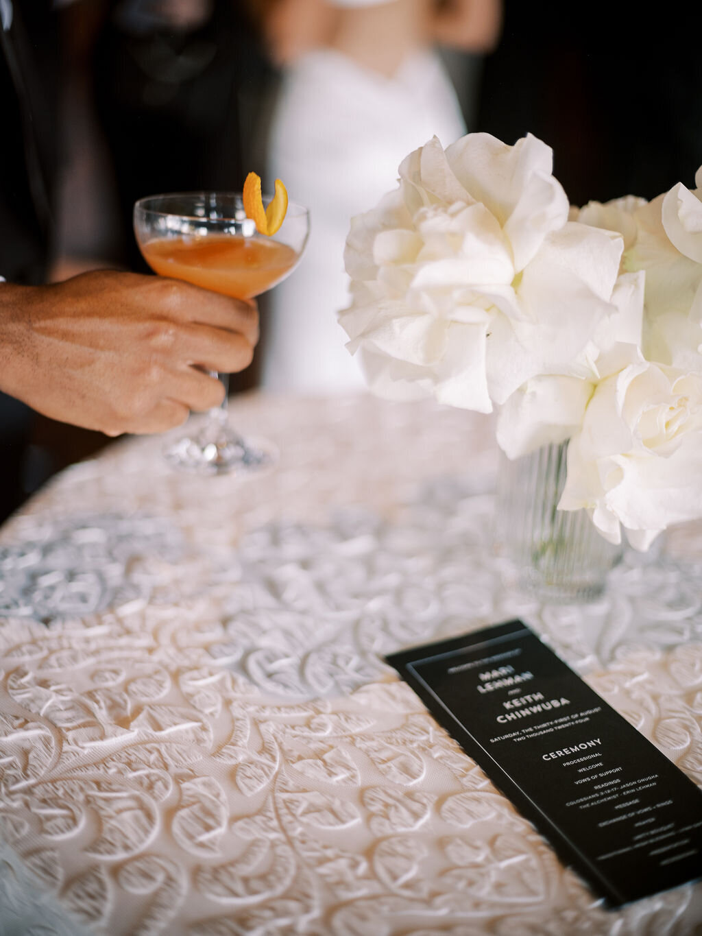 A hand holds a cocktail next to white roses and a black menu card on a textured tablecloth.