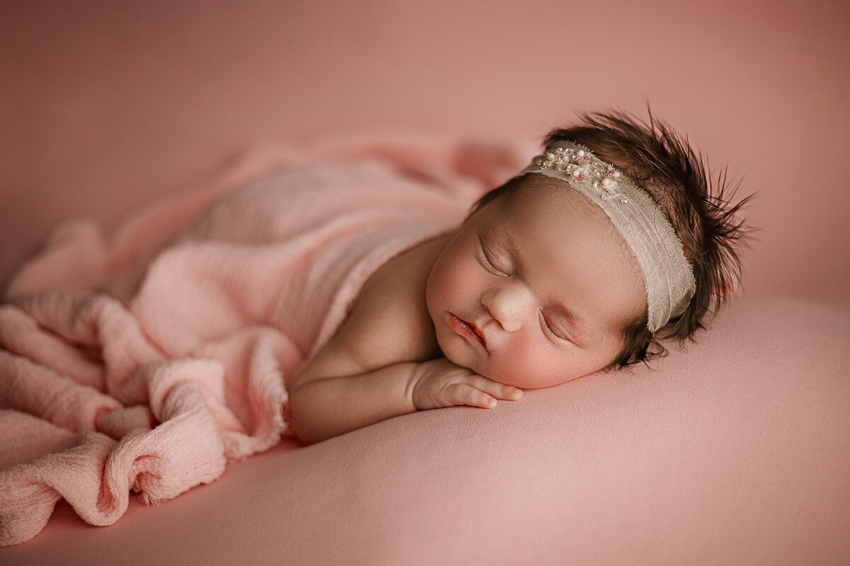 Sleeping newborn baby girl in a soft pink wrap and floral headband, photographed in a Maple Grove photography studio.