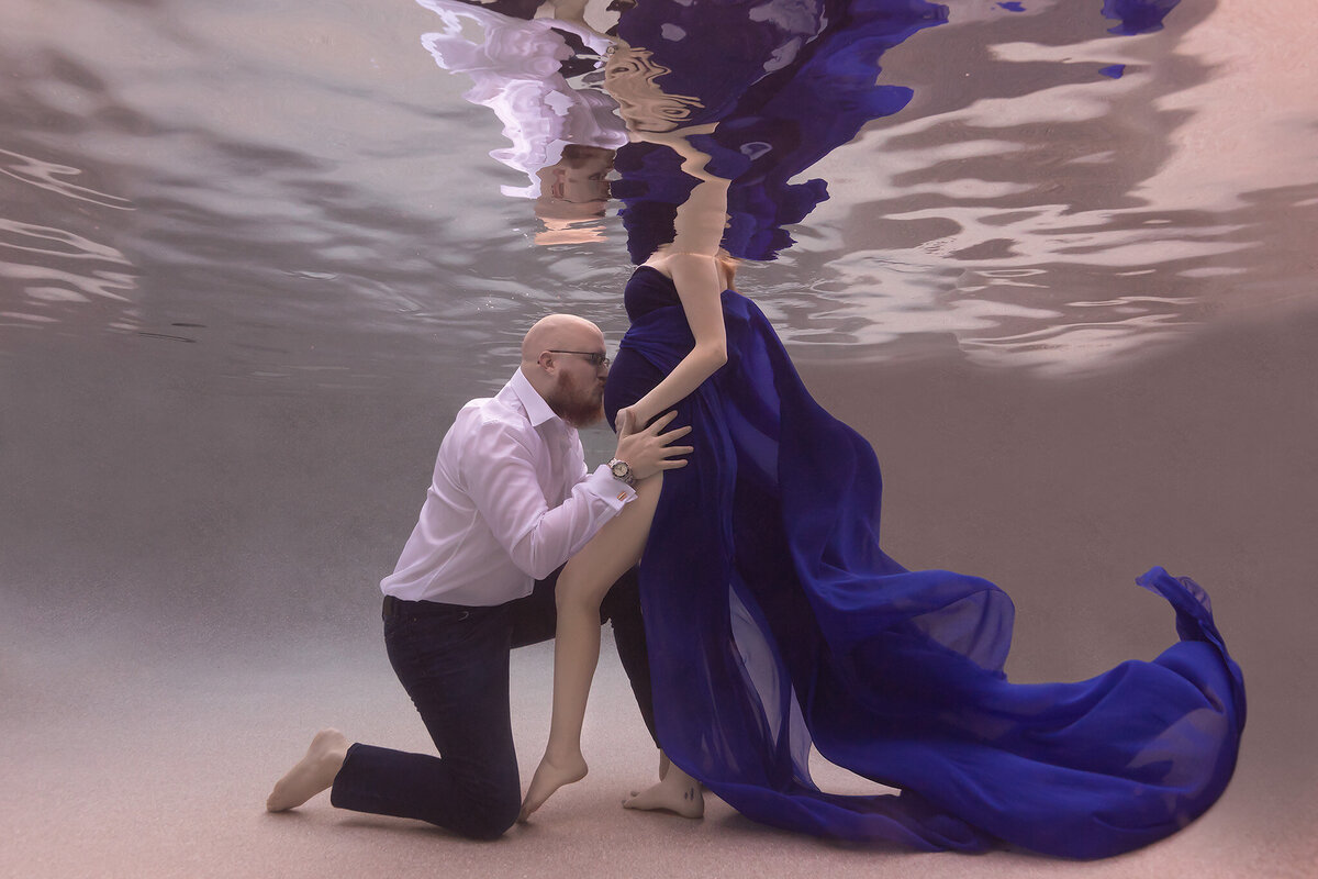 A heartfelt underwater maternity photo captured by a Dallas underwater pregnancy photographer. A man kneels underwater, gently kissing the woman’s belly, cradling her leg with one hand. She wears a flowing blue gown that billows elegantly in the water, creating a striking contrast against the serene, ethereal background. The intimate gesture evokes love, connection, and anticipation.