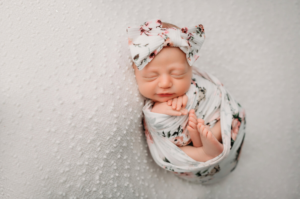 newborn baby girl swaddled in a white floral wrap with flowers on it and big bow on white backdrop
