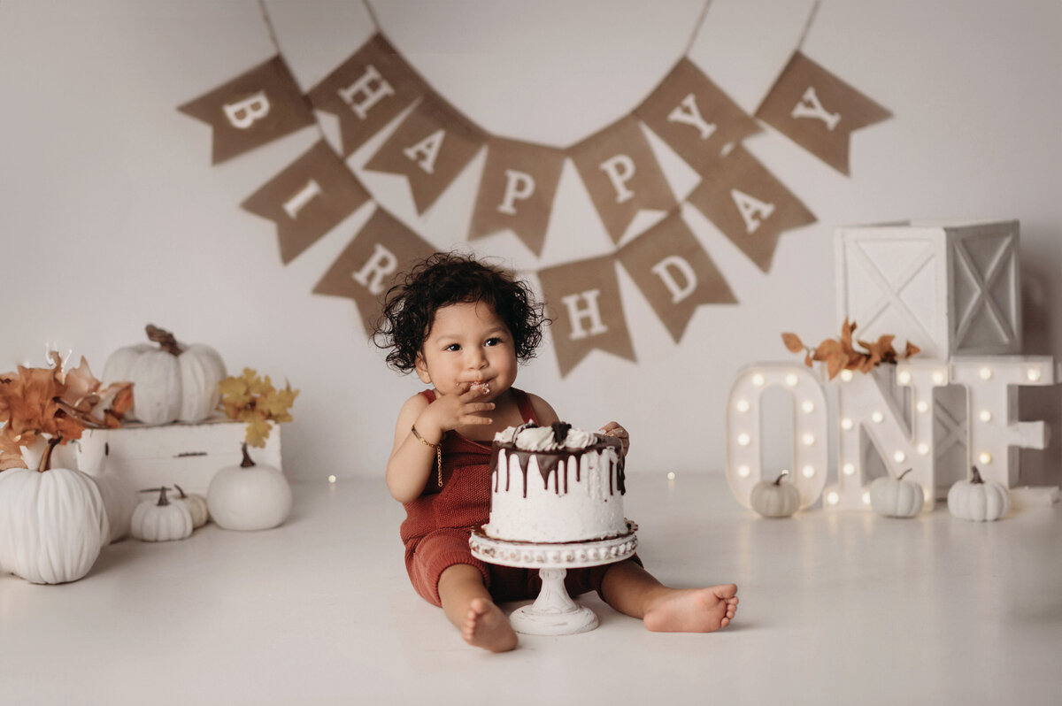 one year old baby boy wearing overalls eating his big white cake and licking his fingers posed in front of  a fall themed backdrop with a happy birthday hanging sign