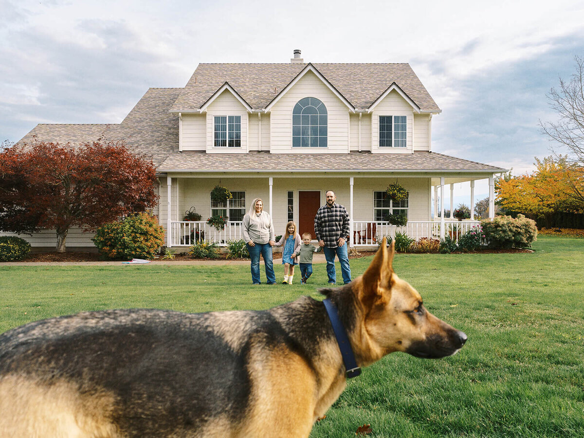 A family stands in front of their house with a dog in the foreground looking away from the camera.