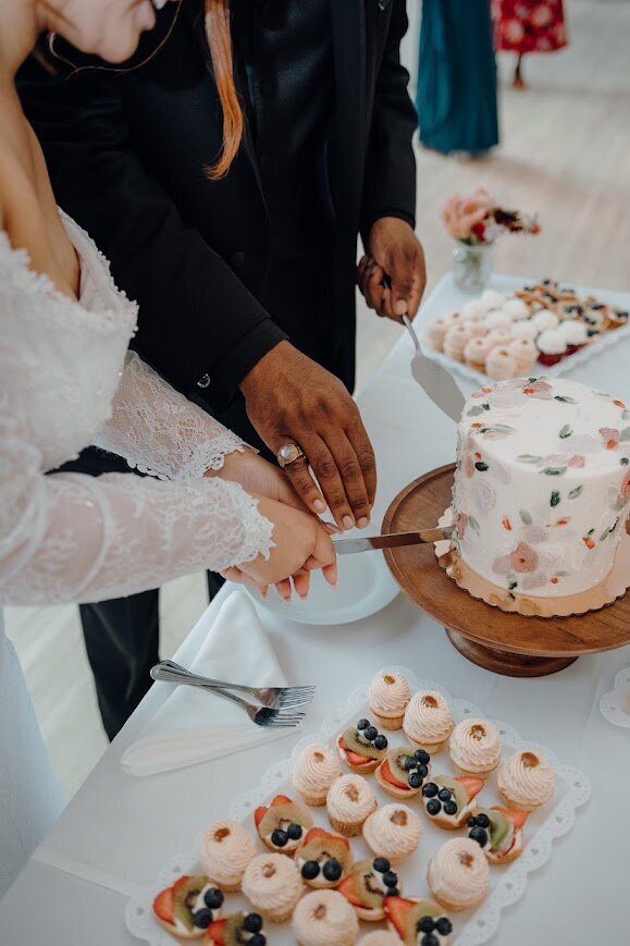Photo shows the hands of a couple in wedding attire, cutting their wedding cake. The cake is white with pallet knife flowers in pinks, white, and red and sits on a wooden stand. Surrounding the cake on the table are other platters with mini cupcakes and fruit tarts.