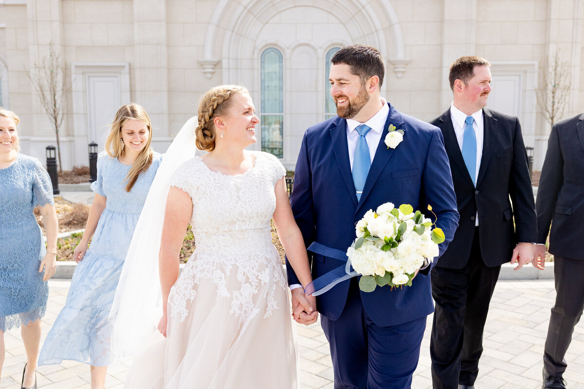 The bride and groom walk hand-in-hand, smiling and looking at each other while their wedding party follows behind.
