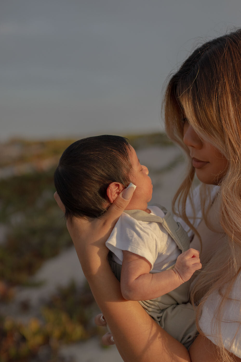 A mother holding her newborn baby close, looking at the child as they stand by the beach