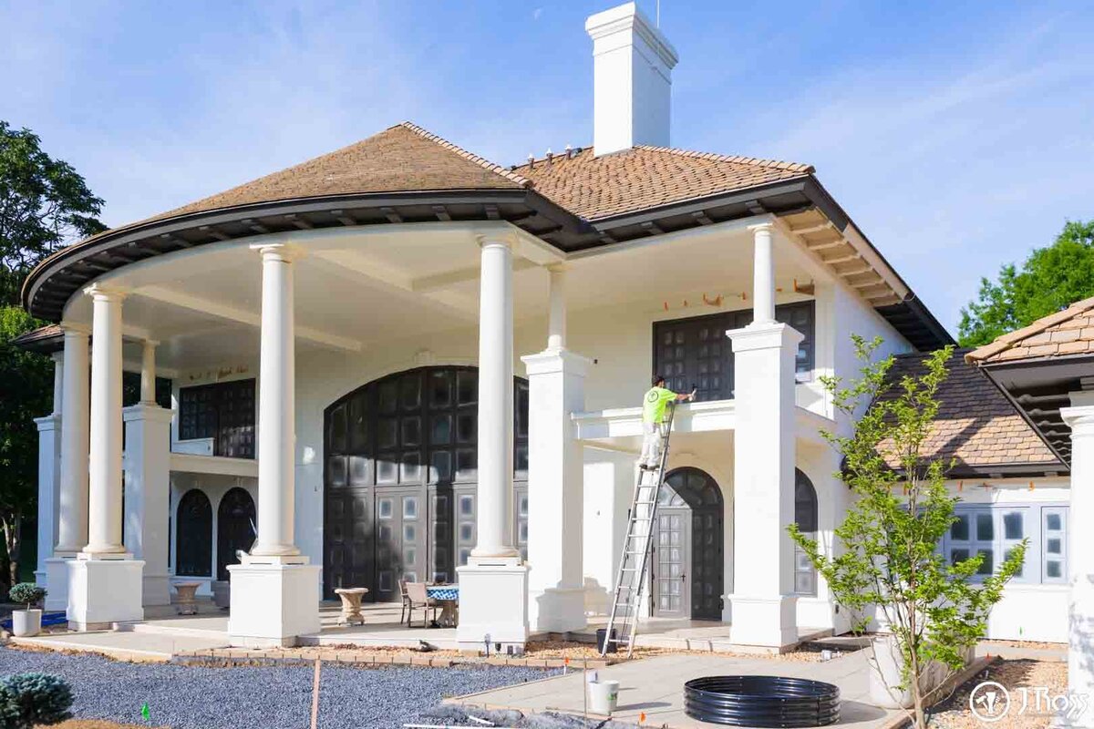 A J.Ross painter on a ladder carefully works on the tall entryway of a stucco mansion with a terracotta roof, showcasing large-scale home exterior painting Bristol, VA expertise.