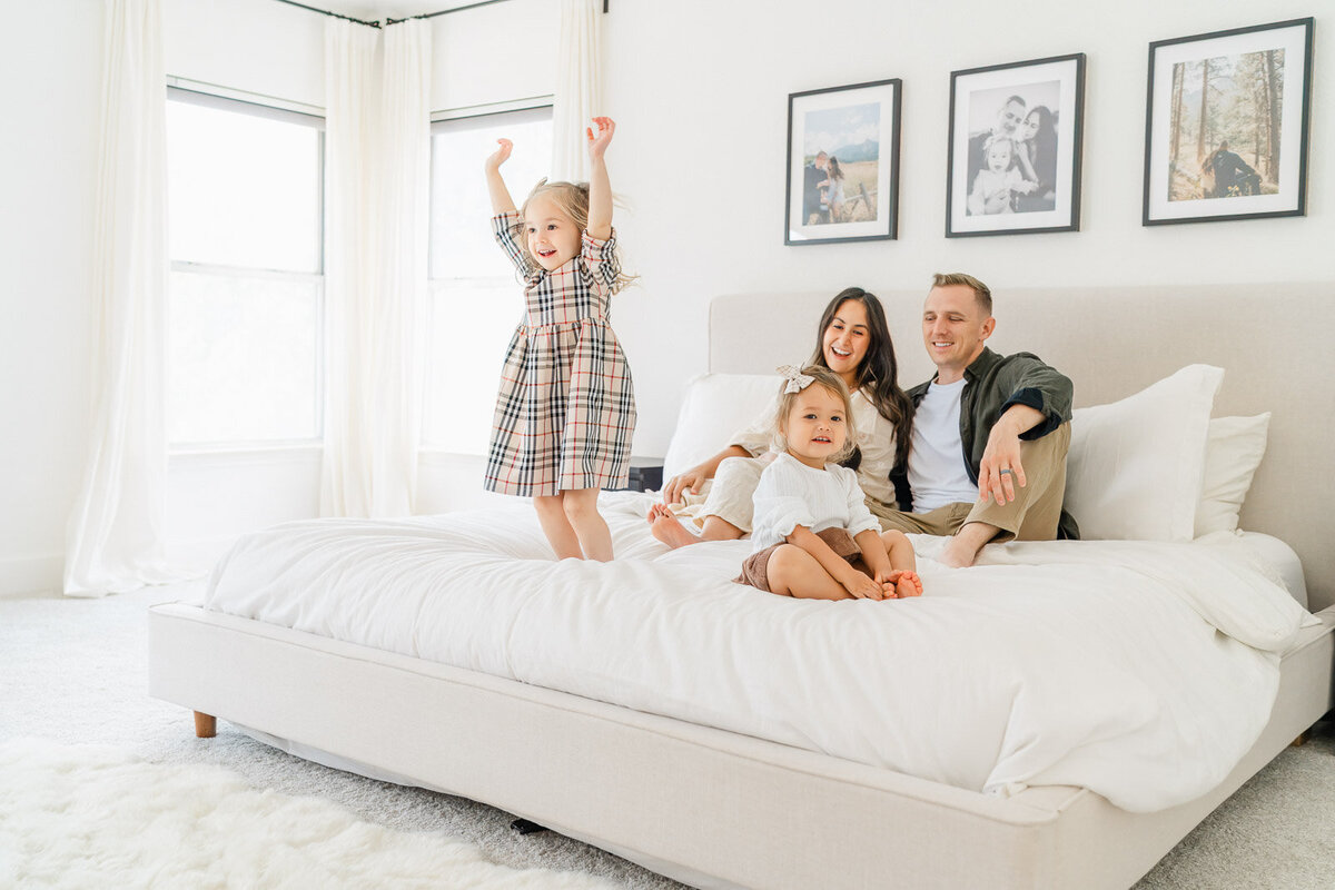 a toddler girl jumps on a bed while her parents hold their newborn in the background and her young sister looks at the camera of their Round Rock newborn photographer.