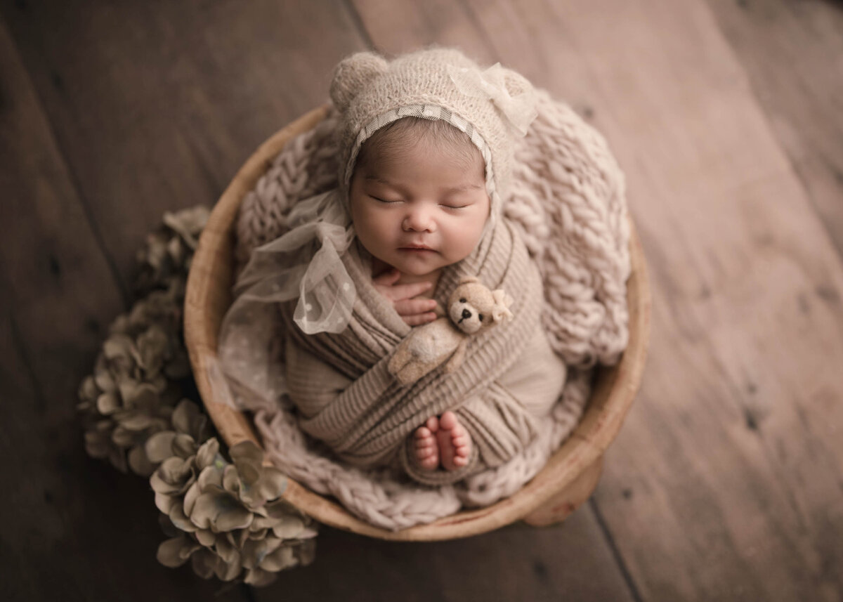 Aerial image. Baby is swaddled in taupe with her fingers and toes peeking out of the wrap. Baby is wearing a beige bonnet with teddy bear ears and has a small felt teddy resting beside her cheek. Captured by best Lake Elsinore newborn photographer Bonny Lynn Photography.