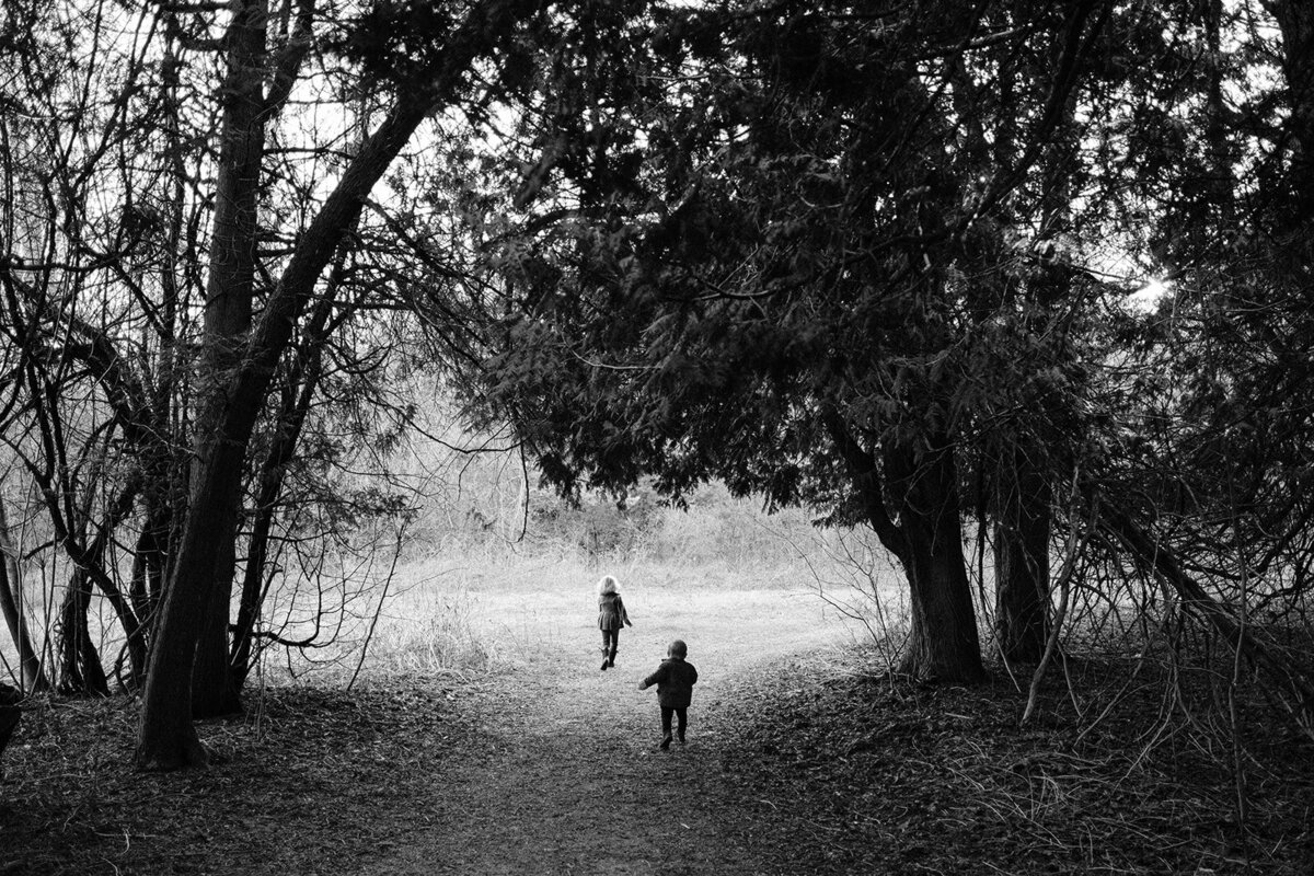 Two children walk on a forest path surrounded by tall trees, captured in black and white by a Toronto photographer.