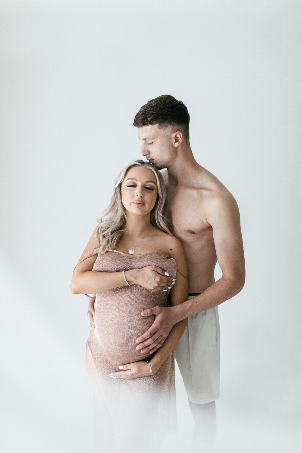 Couple in a white room, the woman is pregnant and the man is holding her belly from behind