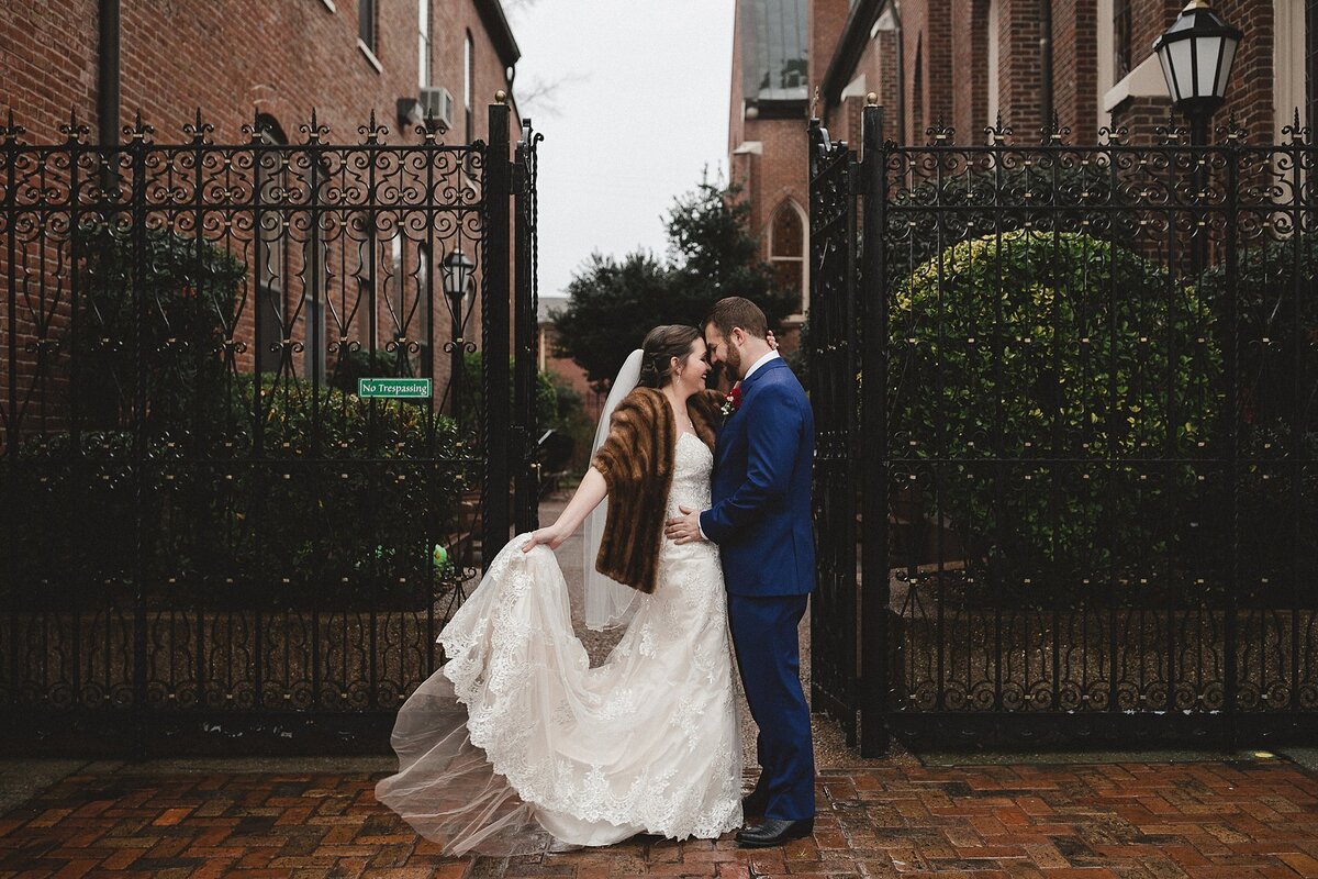 Bride wearing a flowing white lace wedding dress, veil and mink stole kisses the groom wearing a blue suit in front of a Nashville church with a black cast iron gate.