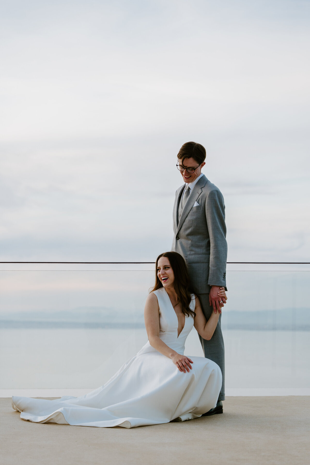 Bride sitting at groom’s feet on modern terrace overlooking water