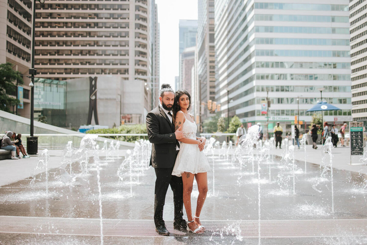 A stylish couple poses confidently in Dilworth Park, surrounded by fountains and city skyscrapers