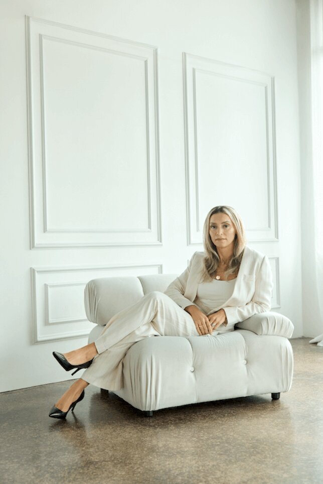 woman in a white suit sitting on a couch with a french wall in the background looking to the camera