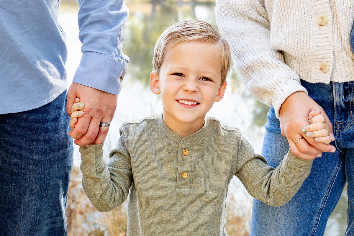 A young boy in a green shirt grins widely while holding his parents’ hands, standing in front of a serene lake