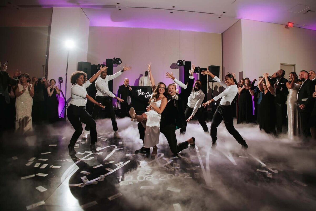 A lively wedding reception dance floor with a couple at the center, surrounded by dancers under dramatic lighting and smoke effects.
