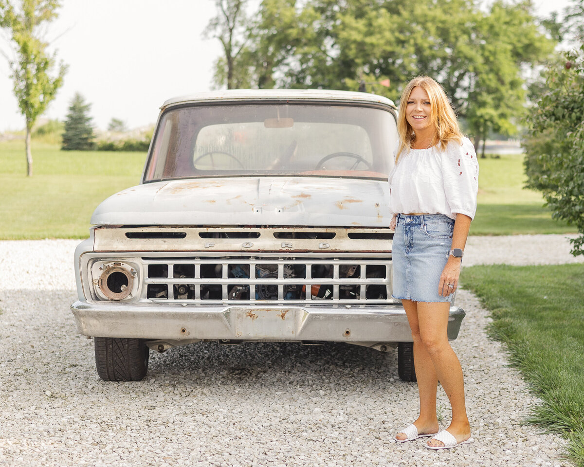 Mom is standing in front of a classic ford truck in a rocked drive, wearing a blue jean skirt, half sleeve white shirt, white sandals, has long blonde hair, smiling at the camera