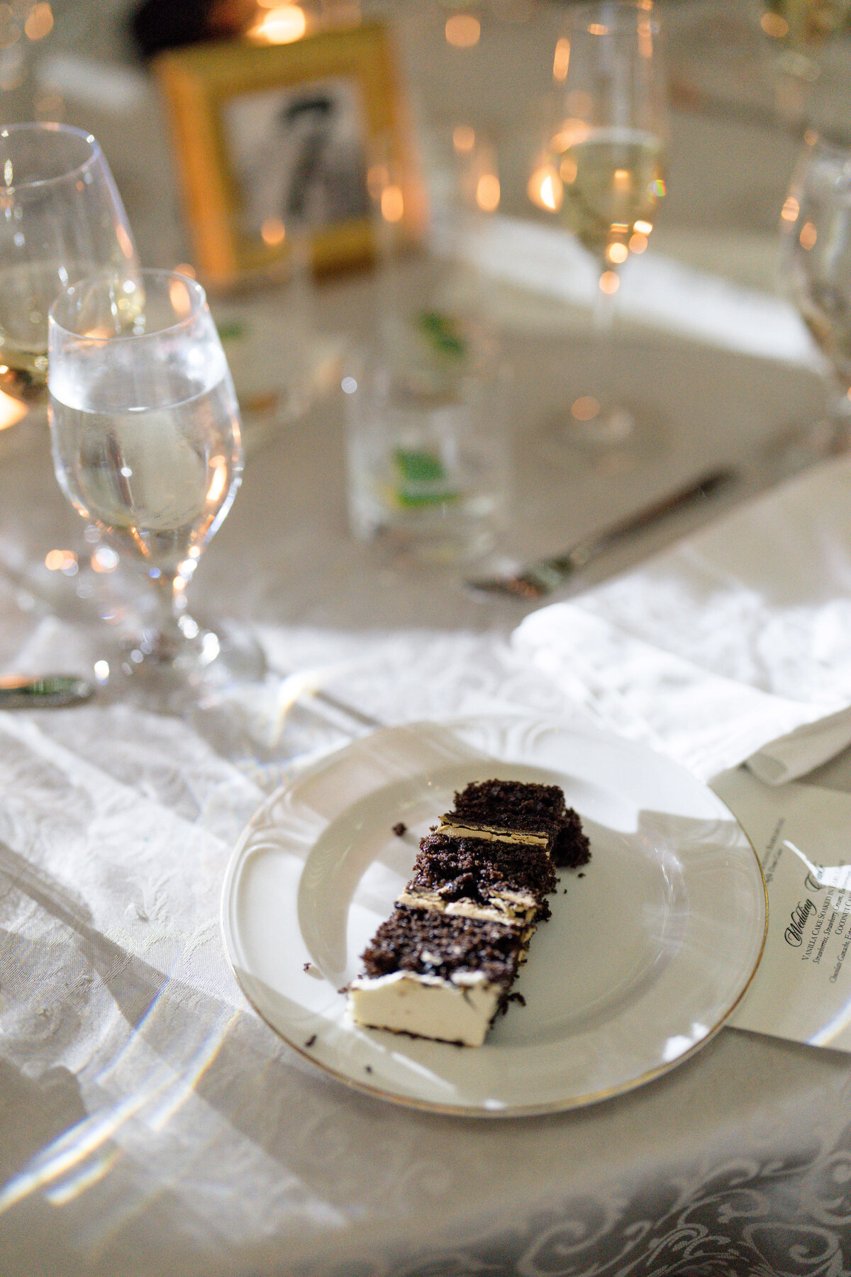 A slice of chocolate and vanilla layer cake with a crumb topping is on a white plate. The table is set with glassware, including wine glasses and water glasses, and theres a blurry photo frame in the background.