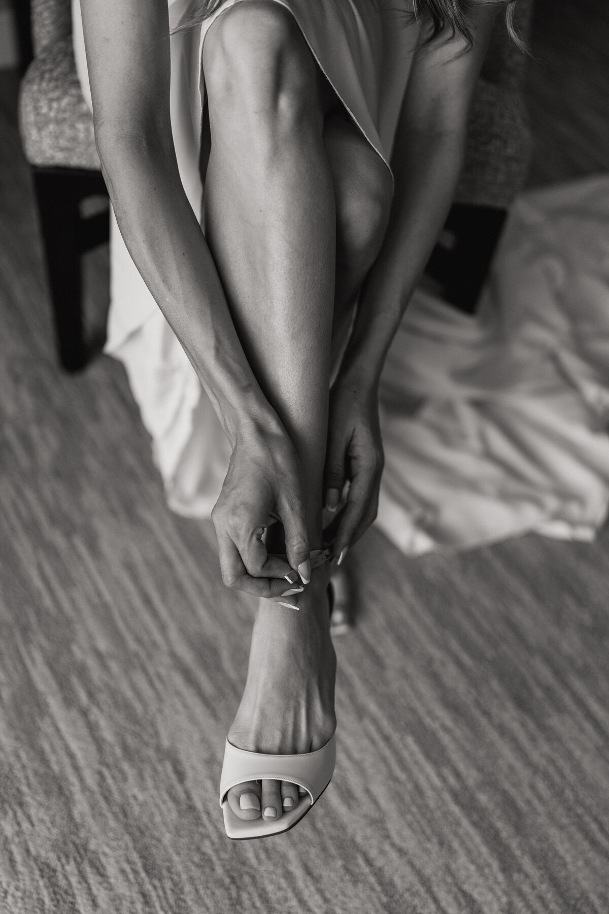 A black and white photograph of Oriel as she gets ready on her wedding day at the Four Seasons Hotel in Westlake Village, California. The photograph is a closeup of the bride's leg, side lit by a window, as she buckles her high heal wedding shoe. Her satin dress is flowing down to the floor behind her. Wedding photography by Stacie McChesney/Vitae Weddings.